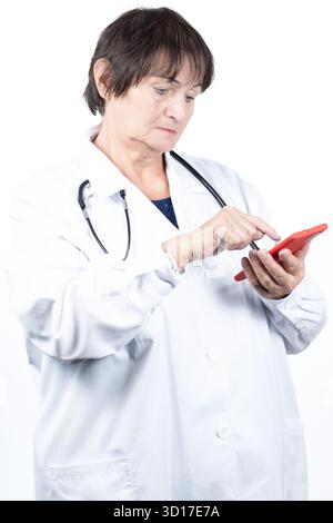 Senior woman doctor wearing a white lab coat and stethoscope, using a red smartphone, representing digital healthcare, online consultations, Stock Photo