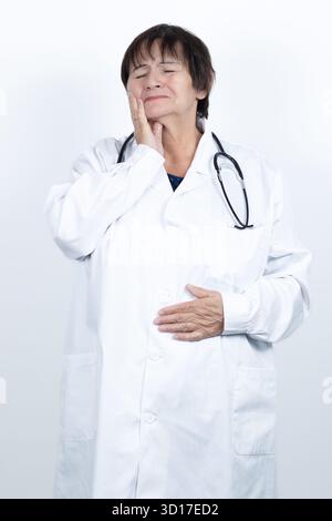 Senior woman doctor wearing a lab coat and stethoscope experiencing severe toothache pain, holding her jaw and closing her eyes in discomfort Stock Photo