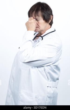 Female doctor in white coat pressing hand to eyes and nose bridge, showing stress, burnout and headache from long hours in healthcare, exhausted Stock Photo
