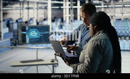Server room coworkers use laptop to install updates and keep AI systems running. Data center engineering team uses notebook to write code improving infrastructure output, camera B Stock Photo