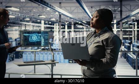 Data center admin monitors deep learning process with laptop, reviewing operational metrics. African american woman using device to review neural networks reports during inspection, camera A Stock Photo