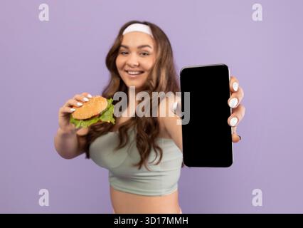 Cheerful Young Plus Size Woman in Sportswear Shows Smartphone While Holding a Burger Stock Photo