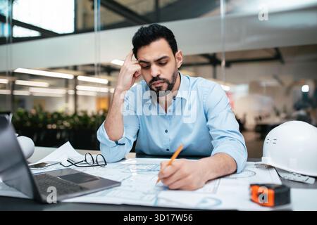 Engineer Focuses on Design Projects While Working in an Office Space, Surrounded by Plans and Tools During a Productive Workday Stock Photo
