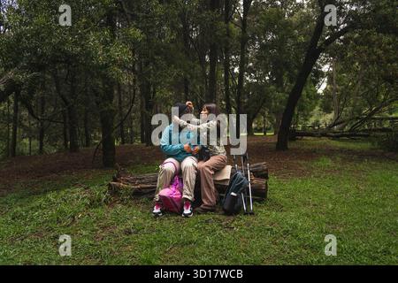 Female couple sitting on a log, exchanging loving gestures in a green forest Stock Photo