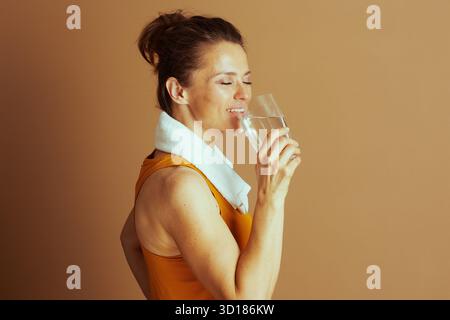 A mid-adult woman with a messy bun smiles peacefully with closed eyes, drinking water from a glass. Wearing an orange tank top and white towel, she em Stock Photo