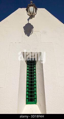 Minimalist architectural detail of a whitewashed wall with a narrow green barred window and a vintage lantern casting a strong shadow under blue sky Stock Photo