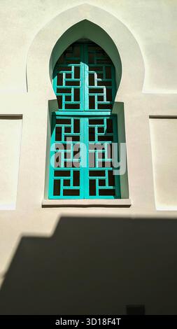 Traditional arched window with geometric turquoise lattice casting shadows on a white stucco wall under bright sunlight Stock Photo