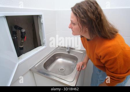 Caucasian woman looking at open access hatch revealing pipes under a sink. Home plumbing maintenance problem and repair concept. Stock Photo