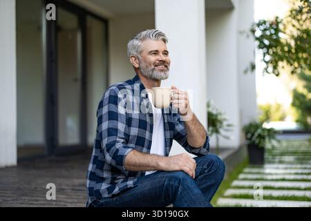 Gray-haired mature man sitting in his house yard and having morning coffee Stock Photo