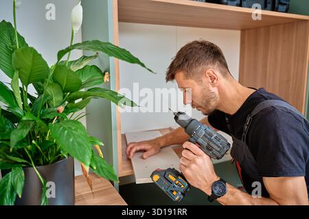 Worker using electric drill to drilling hole in wooden board. Man assembling furniture at home, preparing shelf to installing in self made cabinet. Co Stock Photo