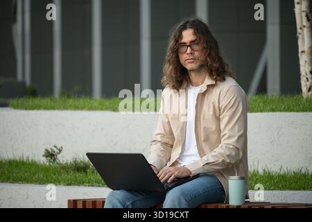 Caucasian businessman or freelancer sits on bench using laptop for remote work at park. Outdoors. Student enjoying work break. Stock Photo