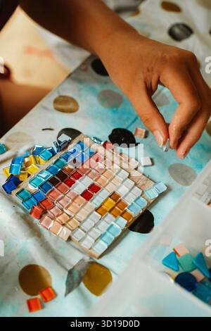 Child carefully places vibrant colored tiles on a mosaic artwork, engaging in a creative arts and crafts activity. Scene shows focus and concentration Stock Photo
