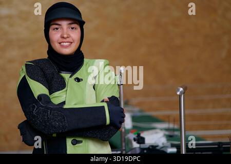 Confident young woman in hijab and shooting gear, smiling indoors, arms crossed, ready for competition, determined athlete, professional sportswoman Stock Photo