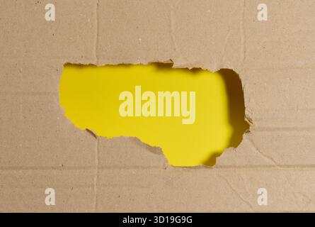 A rough, jagged hole in a sheet of brown corrugated cardboard, revealing a bright yellow background. Copy space Stock Photo