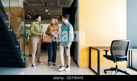 Three young adults interact casually while walking in a contemporary office space, emphasizing collaboration, teamwork, and a vibrant workplace atmosp Stock Photo