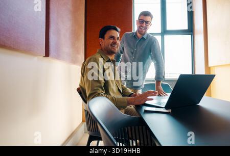 Two mid-adult men are comfortably collaborating at a desk, using a laptop in a brightly lit, modern office space that reflects teamwork and creativity Stock Photo