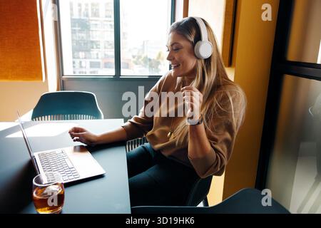 A professional woman enjoys music through headphones while working on her laptop in a bright, modern office. The setting includes natural light and a Stock Photo