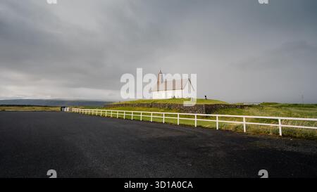 View of a lonely white church standing on a small hill under a cloudy sky, surrounded by a black asphalt road in Reykjavik, Sveitarfélagið Ölfus, Iceland. Stock Photo