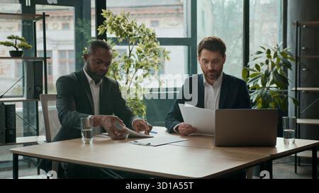 Two diverse smiling business men team businessmen managing documents multiethnic co-workers paperwork cooperate teamwork in office meeting partnership Stock Photo