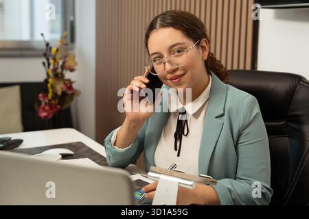 Woman wearing glasses talking on smartphone and working at her desk Stock Photo