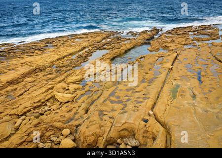 Rocky coast with golden sandstone formations by the sea, small tidal pools, and blue ocean waves under natural light. Stock Photo