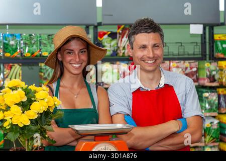 Smiling man and woman working in a greengrocer, standing behind scale and flowers Stock Photo