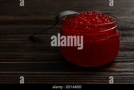 A close-up, studio shot of bright red fish roe (caviar) in a small, clear glass jar with a spoon resting on the rim. Stock Photo