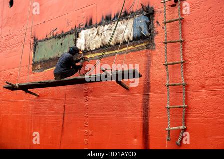 Worker Welding On Suspended Platform Repairing Ship Hull On Red Vessel Exterior Stock Photo