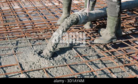 Concrete being poured over steel reinforcement grid. Workers in rubber boots pour wet concrete over a rusty rebar framework during the construction of Stock Photo
