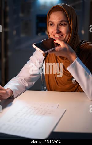 Concept of Artificial intelligence in medicine. Muslim doctor in hijab command to ai assistant, dictate audio message using smartphone working on nigh Stock Photo