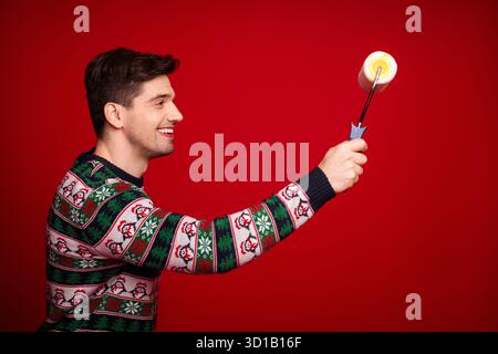 Funny cheerful man in Christmas sweater holds a paint roller against a red background ready to paint holiday magic and fun Stock Photo