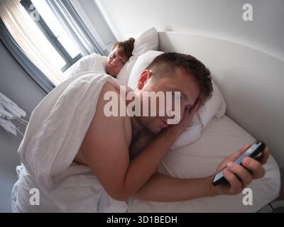 Man using his phone at night while his wife sleeps beside him, bad habit and screen addiction Stock Photo