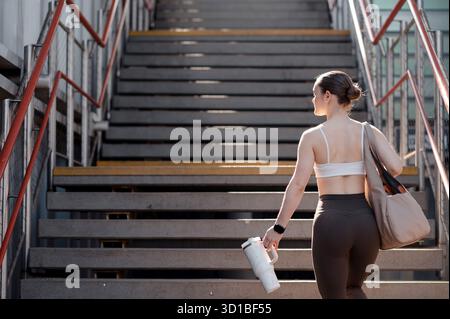 Cute woman walking up stairs carrying a water bottle and a bag, wearing athletic clothing Stock Photo