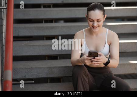 Young woman sitting on stairs, wearing a sports bra and leggings, smiling while using a smartphone Stock Photo