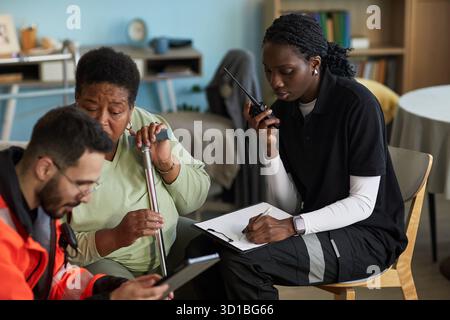 Middle aged Black woman holding cane sitting next to young female nurse using walkie talkie and clipboard with male paramedic showing information on tablet, discussing emergency hospitalization plan Stock Photo