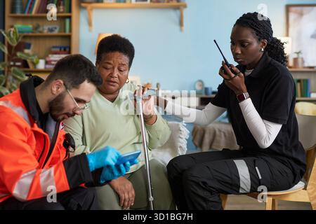 Middle aged Black woman, sitting with cane, receiving assistance from young adult Caucasian man in emergency uniform, while young adult Black woman using walkie talkie nearby Stock Photo