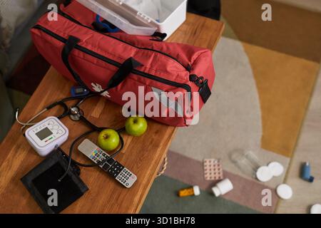 Medical emergency kit with stethoscope, blood pressure monitor, medication blister packs, apples, remote controls and scattered pills on wooden table and carpeted floor during patient health crisis Stock Photo