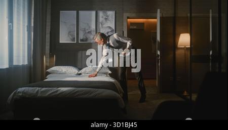 Elegant Mature Hotel Manager in Formal Suit Inspects Luxury Hotel Room, Using Tablet Photograph and Evaluate Bed Arrangement. Concept Hospitality Standards, Quality Control, Professional Management. Stock Photo