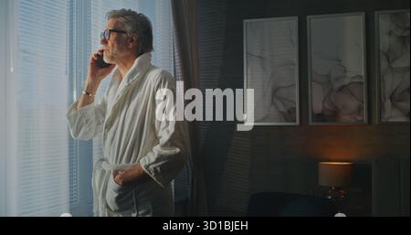 Man in White Hotel Bathrobe Stands by Sunny Window, Holding Phone Call in Hotel Room. Remote Work Lifestyle, Merging of Business and Leisure Travel, and Staying Professionally Connected While on Road. Stock Photo