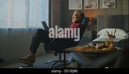 Focused Senior Businessman Works on Laptop in Soft Morning Light of Hotel Room. Breakfast Waits Nearby as Man Stays Deep in Thought. Concept of Remote Work, Travel, and Modern Career. Static Shot. Stock Photo