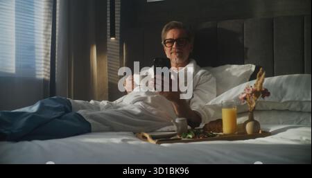 Mature Executive Sits on Hotel Bed, Dressed in Bathrobe, Checking Smartphone While Holding Cup of Coffee For Breakfast. Work Life Integration, Using Mobile Technology, Business Trip. Dolly Shot. Stock Photo