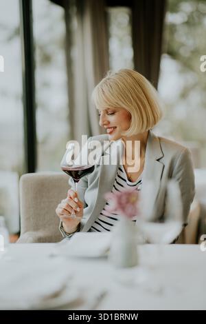 A woman with a short haircut is savoring a glass of red wine at a nicely set restaurant table. She is smiling while engaging in conversation and enjoy Stock Photo
