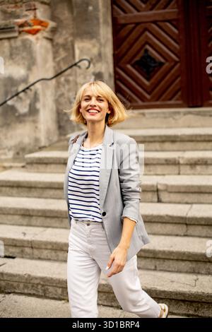 A woman with blonde hair smiles as she walks down stone steps. She wears a casual striped shirt and light pants. The setting features a rustic buildin Stock Photo