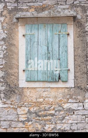 A weathered stone wall frames a faded red inscription above a worn blue wooden shutter. The rustic facade evokes heritage, aging textures, and quiet, Stock Photo