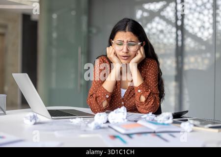 Young woman feeling overwhelmed and frustrated by her work, sitting at a desk with a laptop and crumpled papers, showing signs of stress and a tight deadline Stock Photo