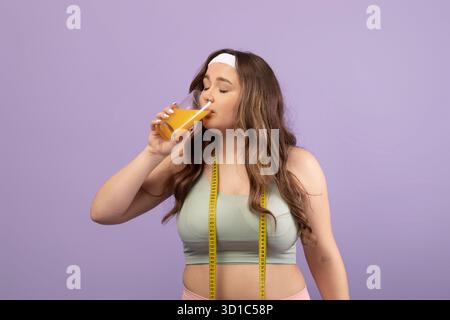 Calm Plus Size Woman Enjoying Orange Juice in Sportswear With Measuring Tape on Violet Background Stock Photo