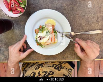 A person is about to cut into a savory breakfast dish of eggs benedict with smoked salmon, hollandaise sauce, and herbs, served on a white plate with Stock Photo