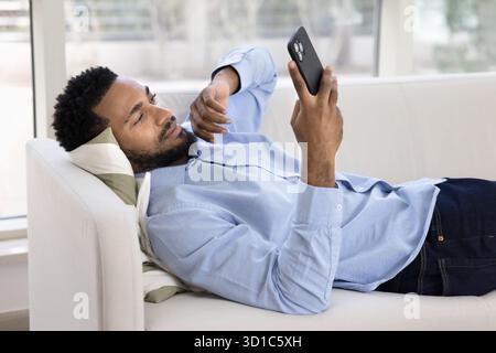African man lying on sofa and using smartphone Stock Photo