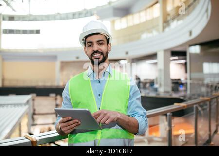 Construction Site Manager in Safety Gear Using a Tablet to Oversee the Project Progress in a Large Indoor Facility Stock Photo