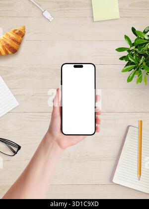 Hand holding smartphone with blank white screen above desk, croissant, notebook, and plant, ready for branding or mobile design Stock Photo
